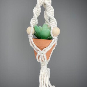 Pode incluir: Um suporte de plantas em macramé creme com um pequeno vaso de terracota com uma suculenta verde. O suporte apresenta contas de madeira e um design com nós, perfeito para adicionar um toque boêmio a qualquer espaço.