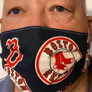 May include: A blue face mask with a white circle containing the words "Boston Red Sox" in red lettering. The circle is surrounded by a red ring. The mask also features a red "B" with a white outline.