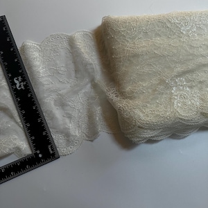 May include: Ivory lace trim with a scalloped edge, partially rolled. A black ruler with white markings is placed on the lace. The lace has a delicate floral pattern. The trim is approximately 3 inches wide.
