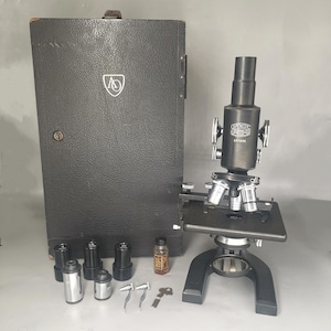 May include: A vintage black microscope with the text "Spencer Buffalo" and a serial number. The microscope is accompanied by a black case, several lenses, and small accessories. The case has a logo on the front.