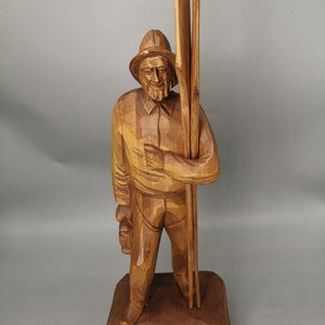 May include: A wooden carving of a man holding two long sticks. The man is wearing a hat and has a beard. The carving is on a wooden base.