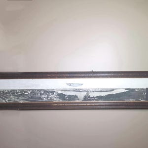 May include: A panoramic black and white photograph of Saskatoon, Western Canada, housed in a dark brown wooden frame. The image displays a wide cityscape with a river, bridge, and railway tracks. The text reads: SASKATOON, SASKATCHEWAN, WESTERN CANADA 1911.