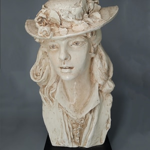 Op de afbeelding: Een wit keramiek buste van een vrouw met een hoed met bloemmotieven. De buste staat op een zwarte basis.