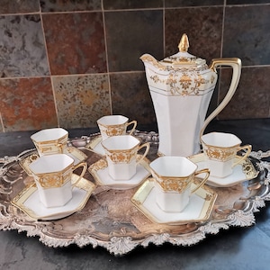 Könnte beinhalten: Ein weißes Porzellan-Tee-Set mit goldenen Akzenten. Das Set beinhaltet eine Teekanne, sechs Tassen und sechs Untertassen. Die Tassen und Untertassen haben eine sechseckige Form. Das Set wird auf einem silbernen Tablett mit verzierten Details präsentiert.