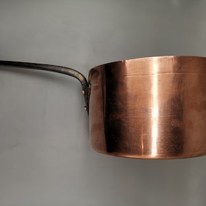 May include: A copper saucepan with a black metal handle. The saucepan is shiny and has a rounded bottom.