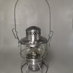 Könnte beinhalten: Eine silberne Metall-Laterne mit einer Glaskugel und einem Drahtgriff. Die Laterne hat ein perforiertes Metalldach und einen Sockel.