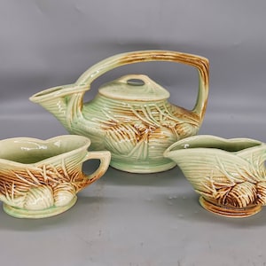 May include: A vintage tea set in a light green and brown colour scheme. The set includes a teapot with a large handle, a creamer, and a sugar bowl. The pieces have a textured, embossed design.