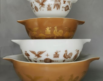 May include: A stack of four vintage Pyrex mixing bowls. Two white bowls with brown illustrations of eagles and wheat, and two amber-colored bowls with gold rooster and wheat designs. The bowls have side handles.