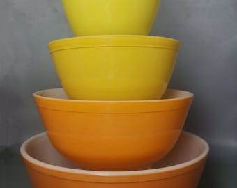 May include: Four vintage mixing bowls in shades of yellow and orange are stacked. The bowls are nested, with the largest at the bottom and the smallest at the top. The two largest bowls have white interiors.