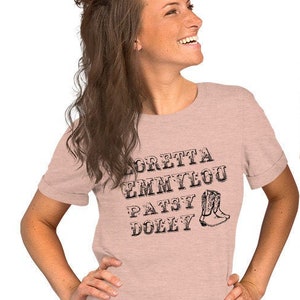 May include: Two people wearing light pink t-shirts with black text that reads "Loretta, Emmylou, Patsy, Dolly" and a black boot graphic.