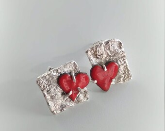 May include: A pair of silver earrings with red heart-shaped charms. The earrings have a textured, rough surface.