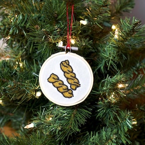 May include: A white fabric ornament with two gold and black beaded twist designs, set in a wooden embroidery hoop. The ornament hangs from a red string against a backdrop of a green Christmas tree.