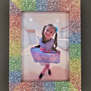 May include: A rainbow glitter picture frame with a photo of a young girl wearing a blue dress and a bow in her hair.