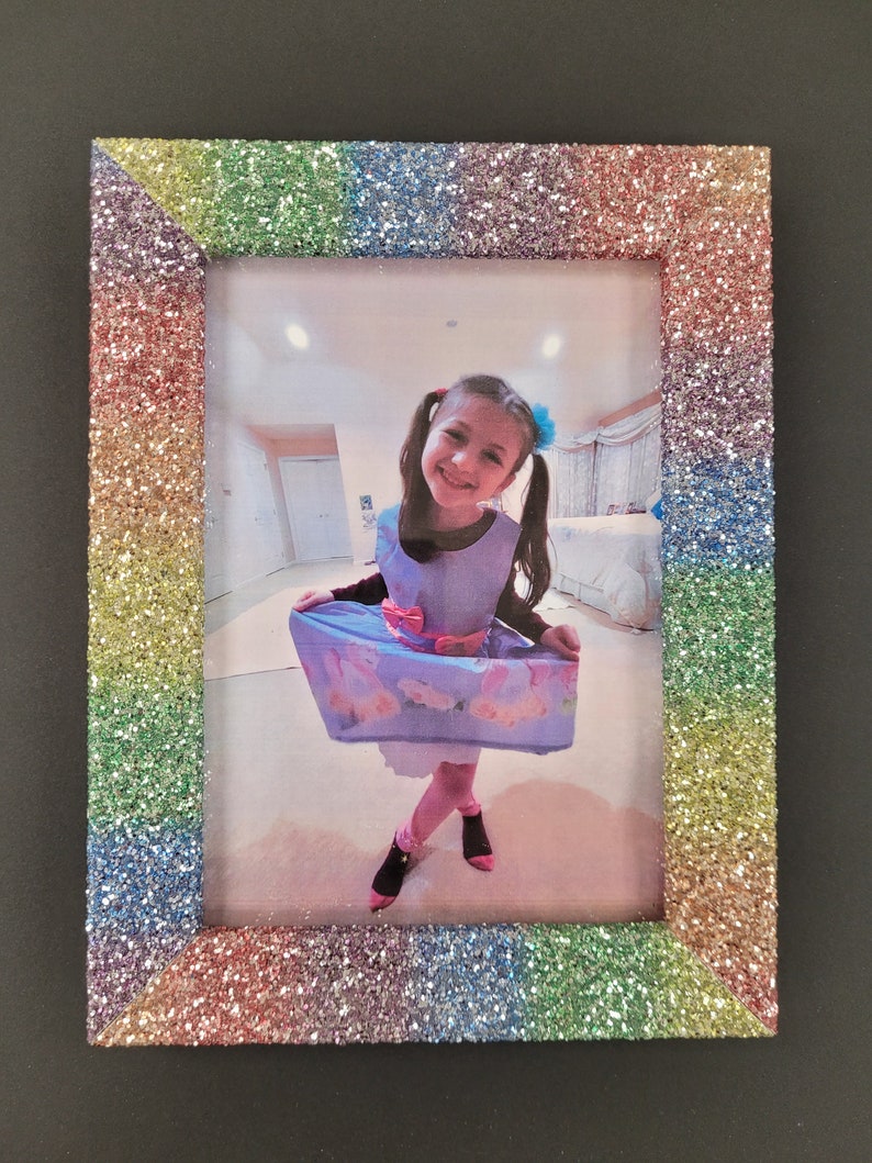 May include: A rainbow glitter picture frame with a photo of a young girl wearing a blue dress and a bow in her hair.