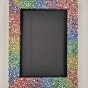 May include: A rainbow glitter picture frame with a black background. The frame is made of cardboard and has a rainbow glitter finish.