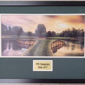 May include: Framed photograph of a golf course with a green fairway, a lake, and a wooden bridge. The sky is a vibrant orange and pink. The text "TPC Sawgrass Hole #17" is on a gold plaque in the bottom right corner.