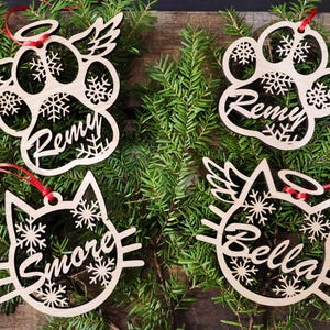 May include: Four wooden Christmas ornaments with snowflake designs and personalized names. The ornaments are shaped like a paw print, a cat, and two paw prints with angel wings. The names on the ornaments are Remy, Bella, Smore, and Remy.