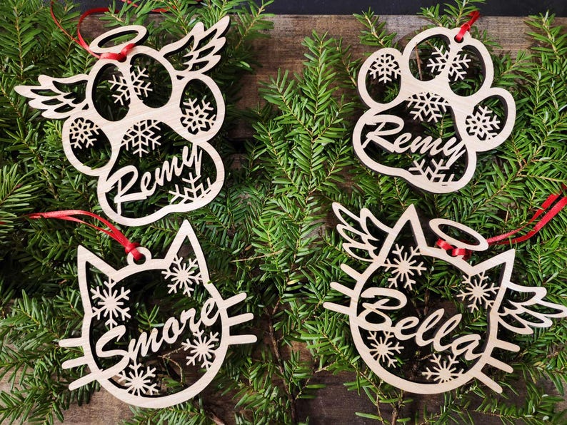 May include: Four wooden Christmas ornaments with snowflake designs and personalized names. The ornaments are shaped like a paw print, a cat, and two paw prints with angel wings. The names on the ornaments are Remy, Bella, Smore, and Remy.