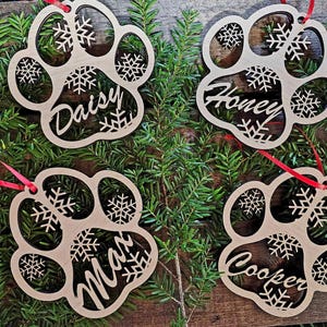 May include: Four wooden paw print ornaments with snowflake designs and personalized names: Daisy, Honey, Max, and Cooper. The ornaments are hanging from red ribbon on a green pine branch background.