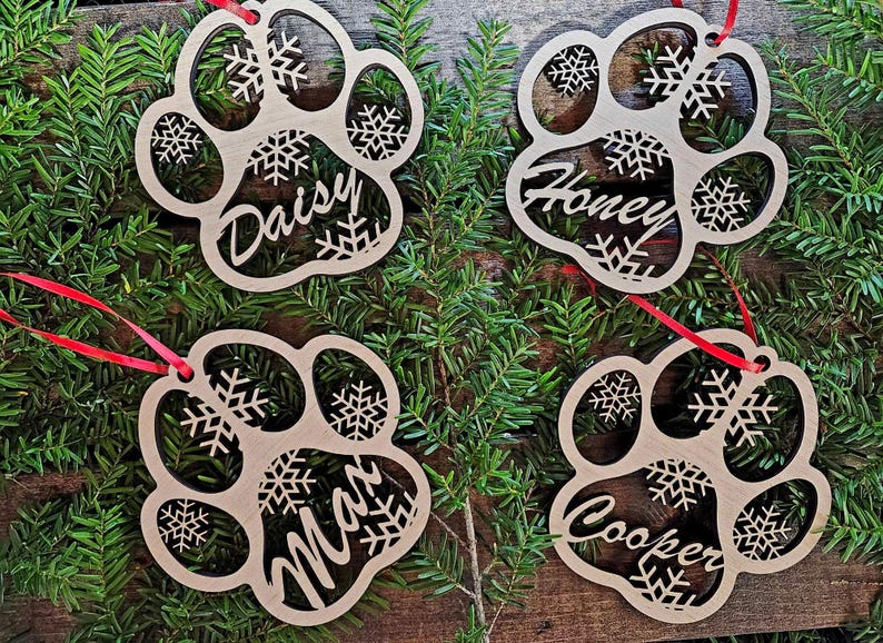 May include: Four wooden paw print ornaments with snowflake designs and personalized names: Daisy, Honey, Max, and Cooper. The ornaments are hanging from red ribbon on a green pine branch background.