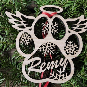 May include: A wooden paw print ornament with angel wings, a halo, and snowflakes. The ornament is personalized with the name "Remy".