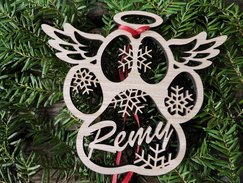 May include: A wooden paw print ornament with angel wings, a halo, and snowflakes. The ornament is personalized with the name "Remy".