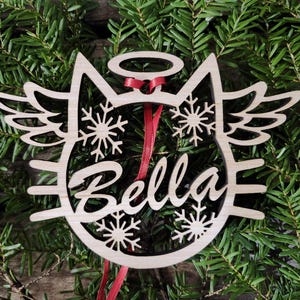 May include: Wooden Christmas ornament shaped like a cat with angel wings, a halo, and snowflakes. The ornament is personalized with the name "Bella".