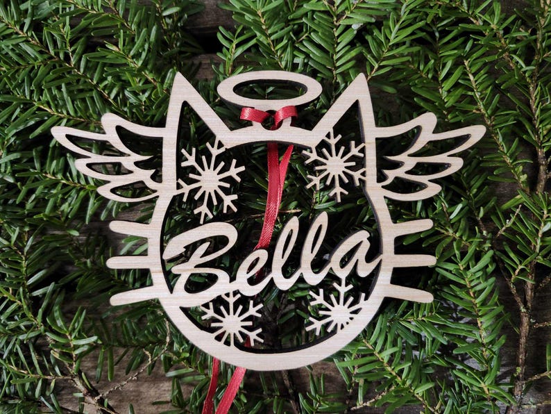 May include: Wooden Christmas ornament shaped like a cat with angel wings, a halo, and snowflakes. The ornament is personalized with the name "Bella".