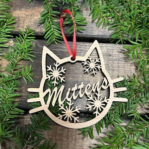 May include: A wooden Christmas ornament shaped like a cat's face with snowflakes and the name "Mittens" cut out. The ornament is hanging from a red ribbon.