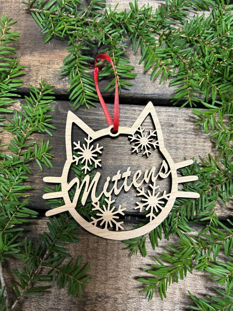 May include: A wooden Christmas ornament shaped like a cat's face with snowflakes and the name "Mittens" cut out. The ornament is hanging from a red ribbon.