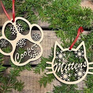 May include: Two wooden Christmas ornaments, one shaped like a paw print with the name "Cooper" and the other shaped like a cat with the name "Mittens". Both ornaments are decorated with snowflakes.