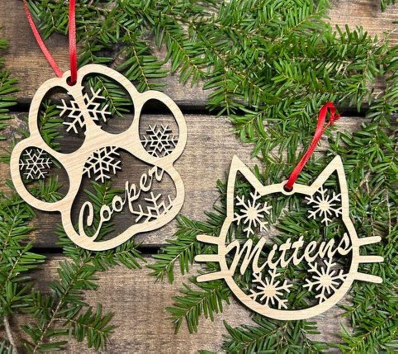May include: Two wooden Christmas ornaments, one shaped like a paw print with the name "Cooper" and the other shaped like a cat with the name "Mittens". Both ornaments are decorated with snowflakes.