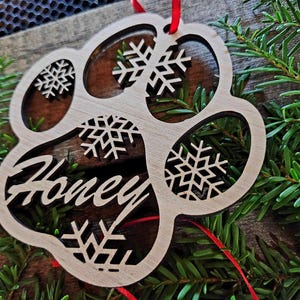 May include: Wooden paw print ornament with a snowflake design and the name "Honey" engraved on it. The ornament is hanging from a red ribbon.