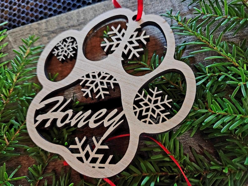 May include: Wooden paw print ornament with a snowflake design and the name "Honey" engraved on it. The ornament is hanging from a red ribbon.