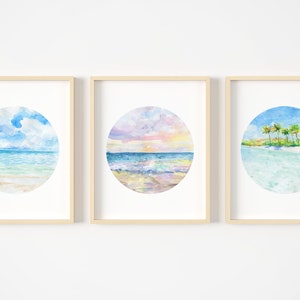 May include: Three framed watercolor paintings of tropical beach scenes. Each painting features a circular image of a beach with blue water, white sand, and palm trees. The first painting shows a cloudy sky, the second shows a sunset, and the third shows a clear sky with palm trees.