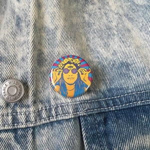 May include: A round button with a colorful image of a person with long hair wearing sunglasses and a flower crown. The background is a blue and red striped pattern.