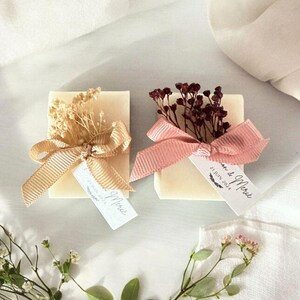 Könnte beinhalten: Zwei quadratische Seifen, jeweils mit Trockenblumen und einem Band verziert. Eine hat ein beiges Band und Blumen, die andere ein rosa Band und dunkelrote Blumen. Jede Seife hat ein Etikett mit der Aufschrift "Merci".