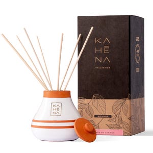 Op de afbeelding: Een witte keramische diffuser met een terracotta top en houten stokjes. De diffuser heeft twee oranje strepen en de tekst "KAHE NA COLLECTION" aan de voorkant. De diffuser staat op een wit oppervlak naast een bruine doos met de tekst "KAHE NA COLLECTION" en "DIFUSOR AROMA DE VERANO" aan de voorkant.