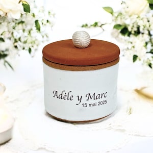 Peut inclure: Un pot en céramique blanche avec un couvercle en terre cuite et un bouton argenté. Le pot est inscrit avec le texte "Adèle y Marc 15 mai 2025".