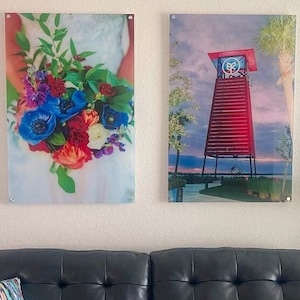 Op de afbeelding: Een zwarte gecapitonneerde leren bank met twee kleurrijke gestreepte sierkussens, geplaatst onder twee grote rechthoekige wandkunstprints, een met een levendig bloemenboeket en de andere met een rode strandwachttoren bij zonsondergang.