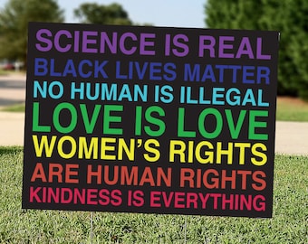 May include: A black yard sign with rainbow coloured text that reads "Science is real, Black Lives Matter, No Human is Illegal, Love is Love, Women's Rights are Human Rights, Kindness is Everything."