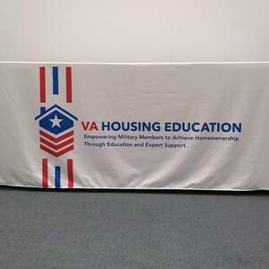 May include: White table cover with a red, white, and blue logo that says "VA HOUSING EDUCATION" and "Empowering Military Members to Achieve Homeownership Through Education and Expert Support."