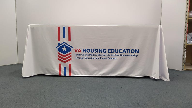 May include: White table cover with a red, white, and blue logo that says "VA HOUSING EDUCATION" and "Empowering Military Members to Achieve Homeownership Through Education and Expert Support."