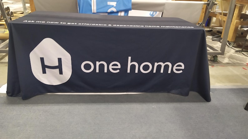 May include: A dark blue table cover with the text "Ask me how to get affordable & accessible home maintenance" and the logo "One Home" with a white house icon and the letter "H" inside.