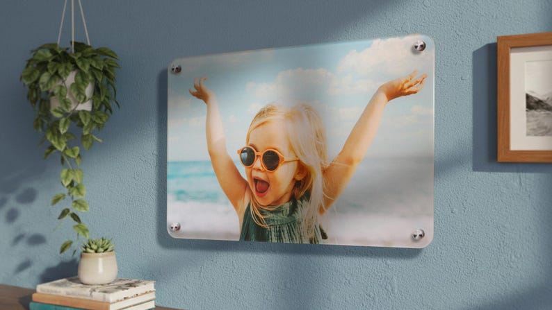 Op de afbeelding: Een rechthoekige acryl wandprint met een foto van een kind met blond haar en een ronde zonnebril, met de armen enthousiast omhoog tegen een strandachtergrond. Bevestigd met vier zilverkleurige metalen afstandhouders.