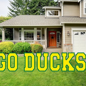 May include: A set of large, yellow, foam letters spelling "GO DUCKS" on a green lawn in front of a house. The letters have a green outline.