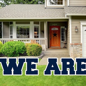 May include: A blue and white yard sign with the text "WE ARE" in large, bold letters. The sign is displayed on a green lawn in front of a house.