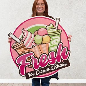 May include: A pink and green round sign with the text "Fresh Ice Cream & Shake" in pink lettering. The sign features a cartoon illustration of ice cream cones and a milkshake.