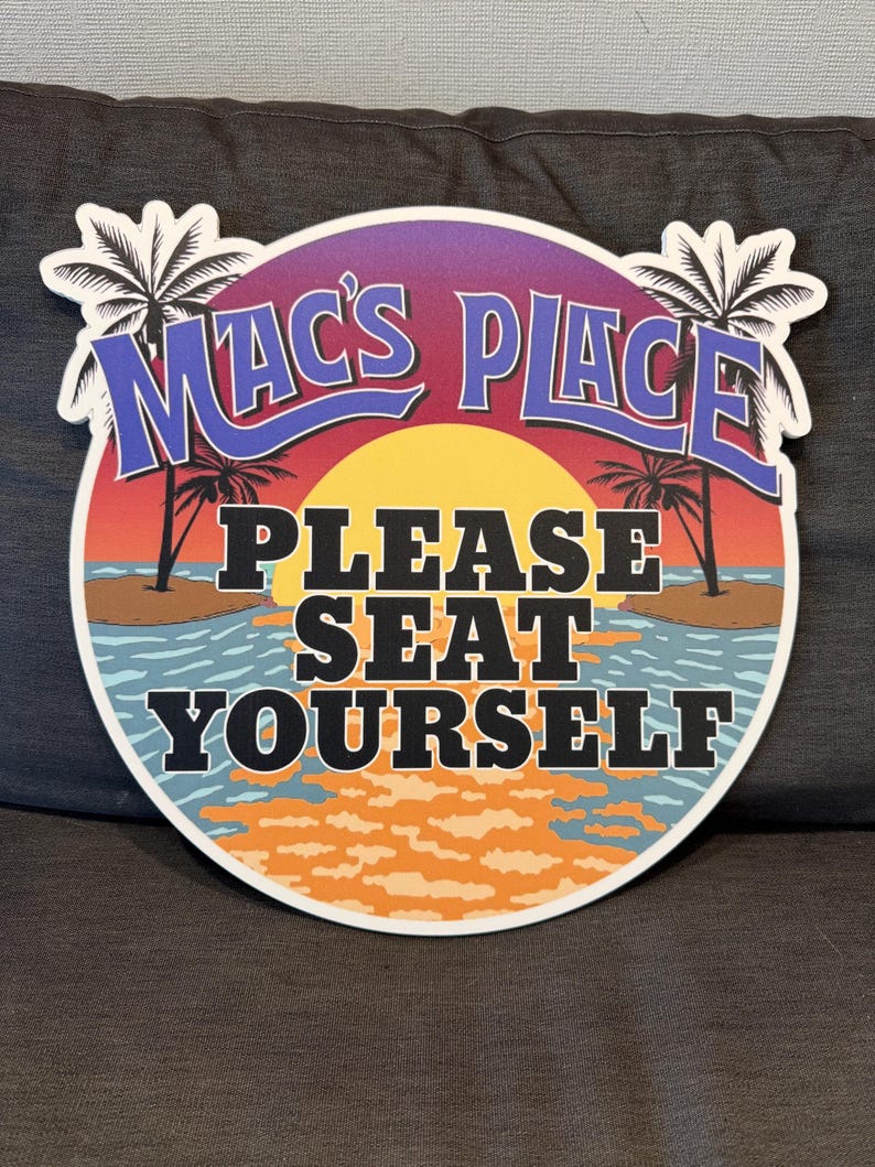 May include: A round sign with a sunset and palm trees. The sign says "Mac's Place" in a blue cursive font and "Please Seat Yourself" in black block letters.