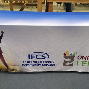 May include: A white tablecloth with a blue gradient at the top. The tablecloth features a superhero child, the text "IFCS Integrated Family Community Services", and the text "ONE CAN FEED" with a graphic of a hand holding a grocery bag.